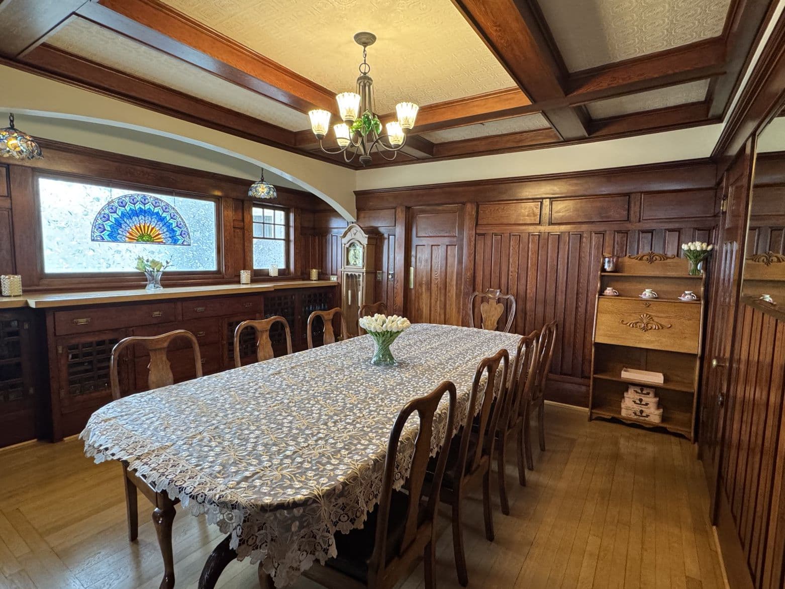 A warm, wooden dining room featuring a long table with a lace tablecloth, stained glass window, and vintage details.