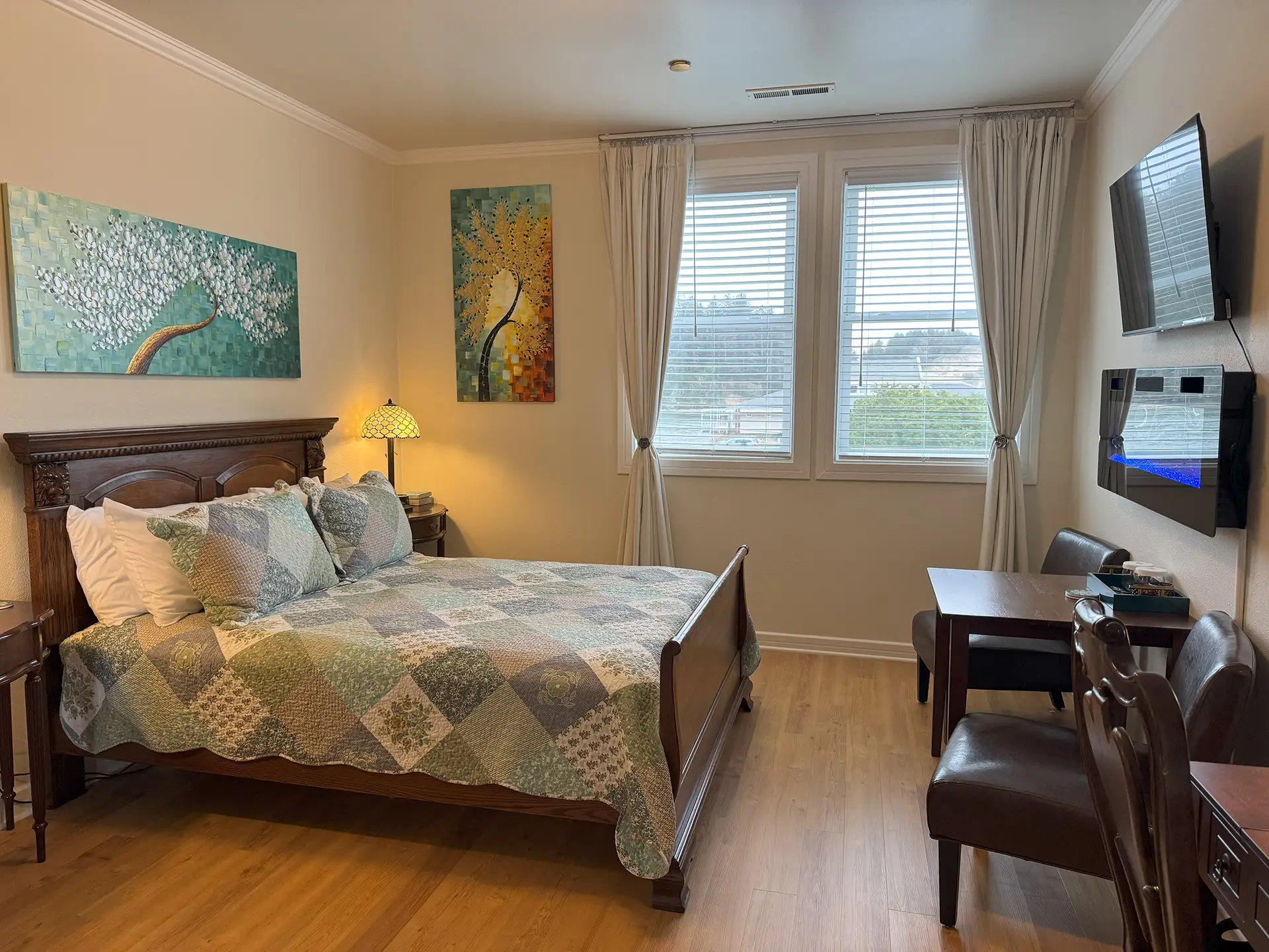A cozy bedroom featuring a quilted bed, two decorative paintings, a lamp, a television, and a small dining table with chairs by the window.