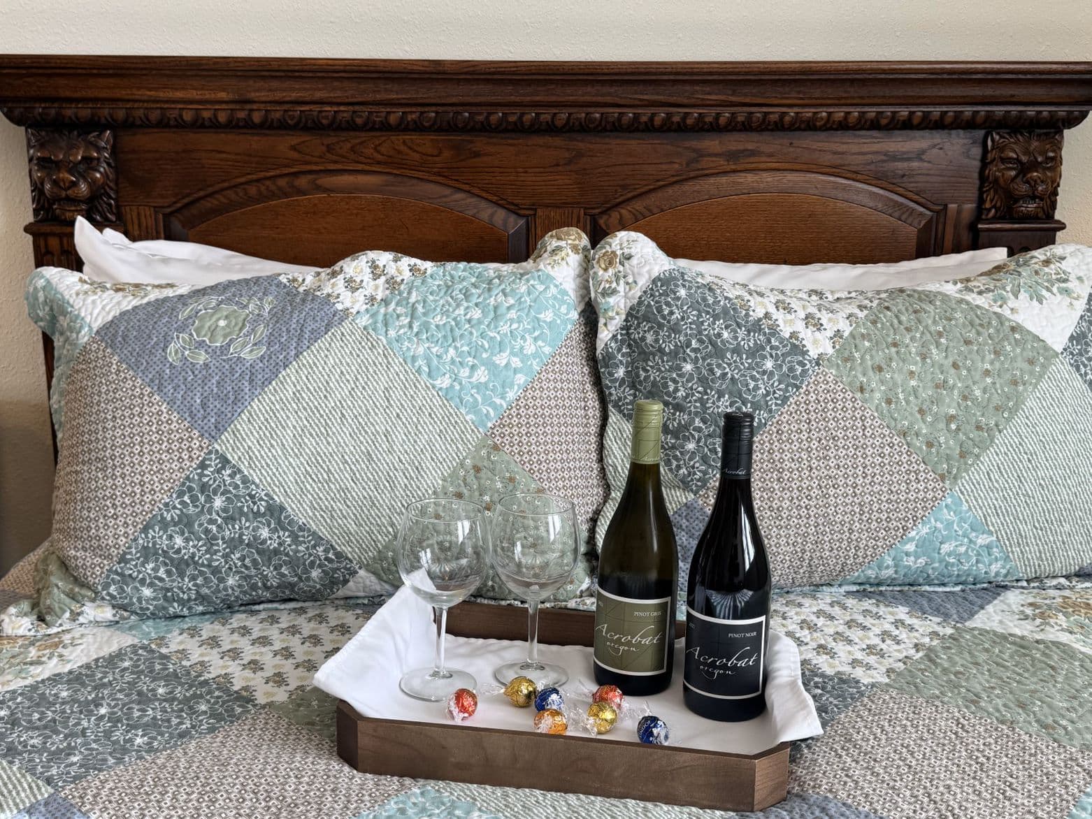 A cozy bed with a colorful quilt, two wine bottles, glasses, and chocolates on a tray.