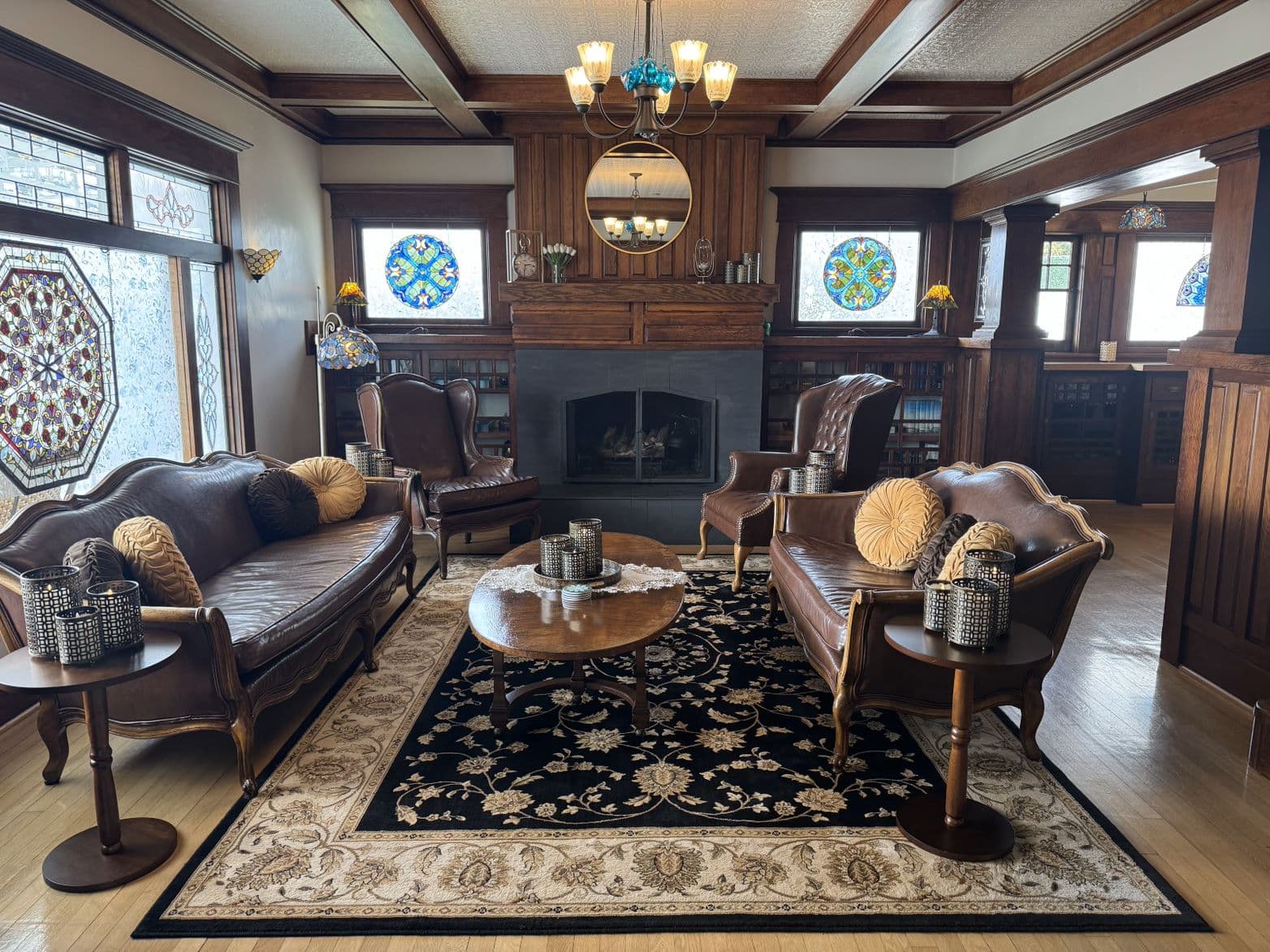 A cozy living room featuring vintage wooden furniture, stained glass windows, and a central fireplace.